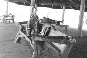 Black and white photo of two peope resting under a covered pavilion. One is lying on a table with their boots off, the other is sitting knees up on a bench completey covered by a sarong apart from their boots.