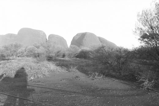 Black and white photo of Kata Tjuta at sunset. There is a shasow of the photographer in the foreground.
