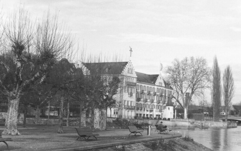 Black and white photograph of a park and buildings along the edge of lake, a man is sitting on park bench