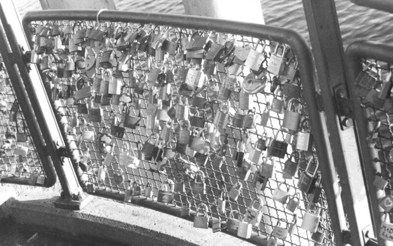 Black and white photograph of a fence covered in love locks (padlocks with names and initials inscribed).