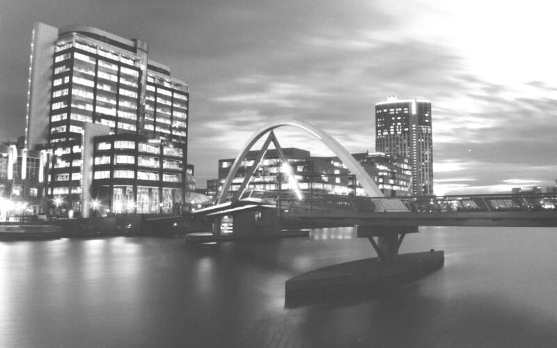 Black and white photo taken at night of a pedestrian bridge spanning a river. The bridge has an arch over the top and is lit up. The surrounding buildings across the water are also lit up.