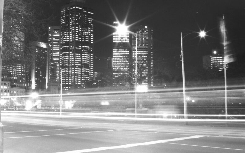 Black and white photo of a city street taken at night. There is a streak of light across the image from passing cars. Skyscrapers are in the background.