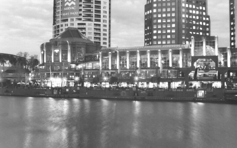 Black and white photo across a river to a promenade of brightly lit buildings and outdoor eating areas.