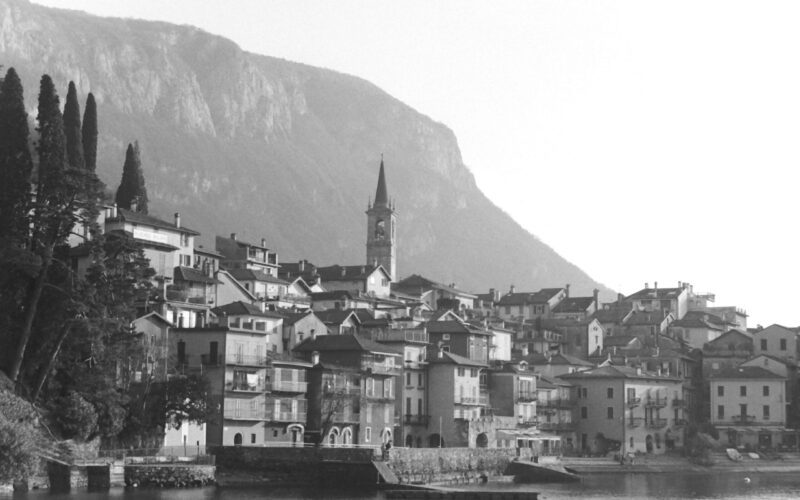 Black and white photo of a lake side village with a clock tower and mountains in the background.