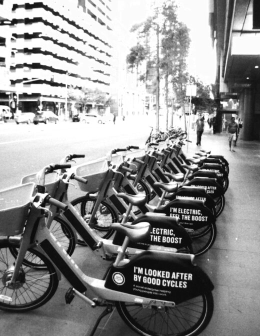 Black and white photo of a row of share bikes parked on a city footpath. The bikes have signs that say "I'm looked after by good cycles" and "I'm electric, feel the boost".