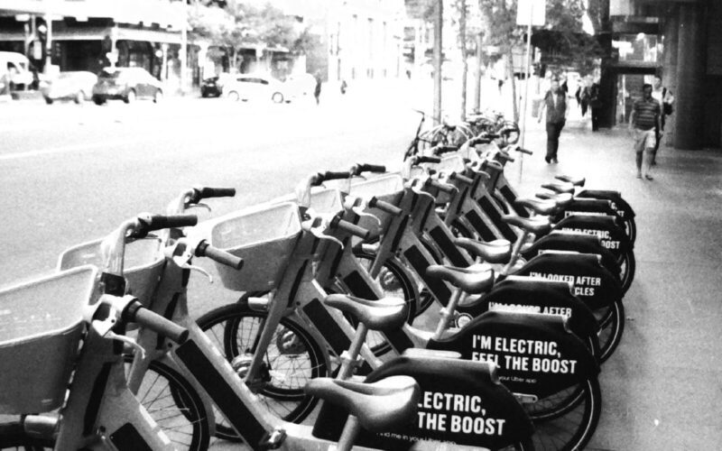 Black and white photo of a row of share bikes parked on a city footpath. The bikes have signs that say "I'm looked after by good cycles" and "I'm electric, feel the boost".