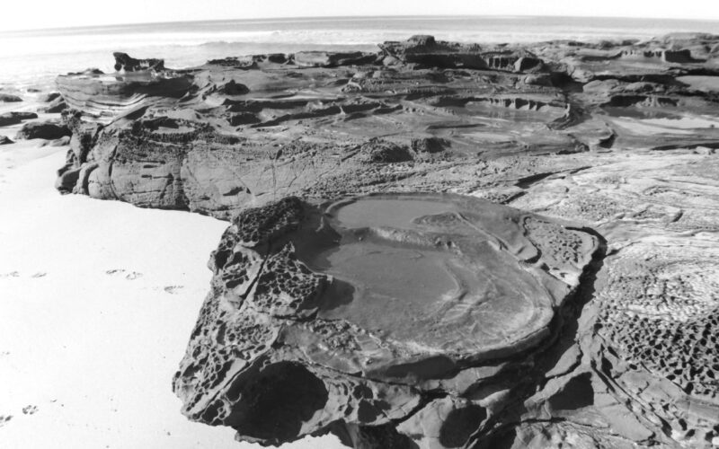 Black and white photo of a large flat rock formation on a beach. The top of the rock contains circular erosion patterns that form many rock pools.