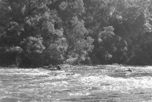 Black and white photograph of two men kayaking down a river towards the camera. The sun is glistening off the water.
