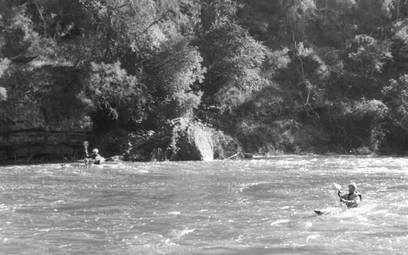 Black and white photograph of two kayaks on a river. There is a man paddling in each kayak and the sun is glistening off the water.