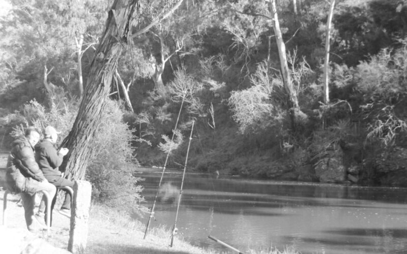 Black and white photograph of two men sitting beside a river with two fishing poles standing in front of them and lines leading into the water. One of the men is looking at the camera.