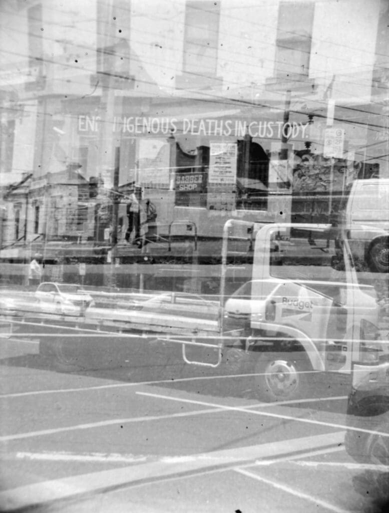 Black and white photo consisting of three exposures. There are multiple vehic;es crossing through the image in front of a building with a sign reading "End Indigenous Deaths in Custody".