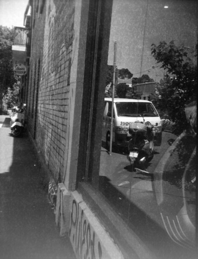 Black and white photo of a Vespa parked on the footpath next to a building, there is another Vespa reflected in the window of the building.