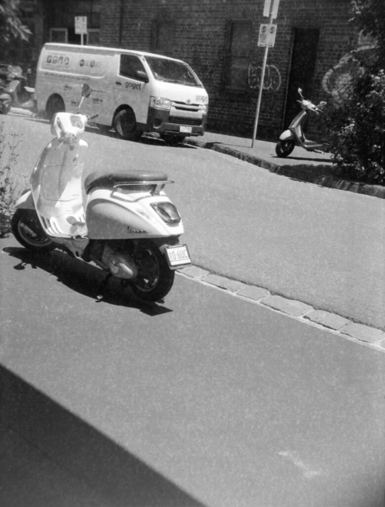 Black and white photo of three vespas parked either side of and across the road from a van with "goget" on the side.