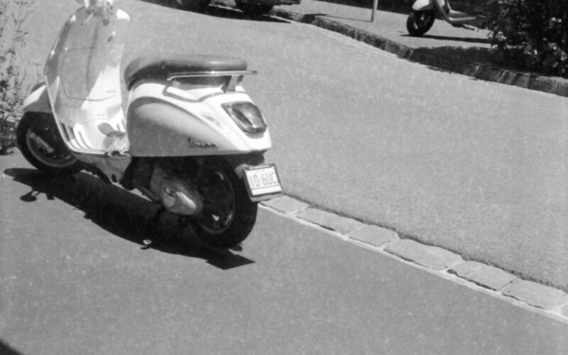 Black and white photo of three vespas parked either side of and across the road from a van with "goget" on the side.