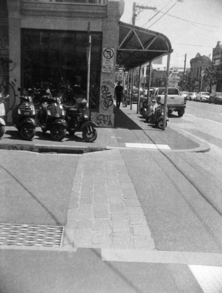 Black and white photo of Vespas lining either side of store on the foot path with a person walking towards the camera