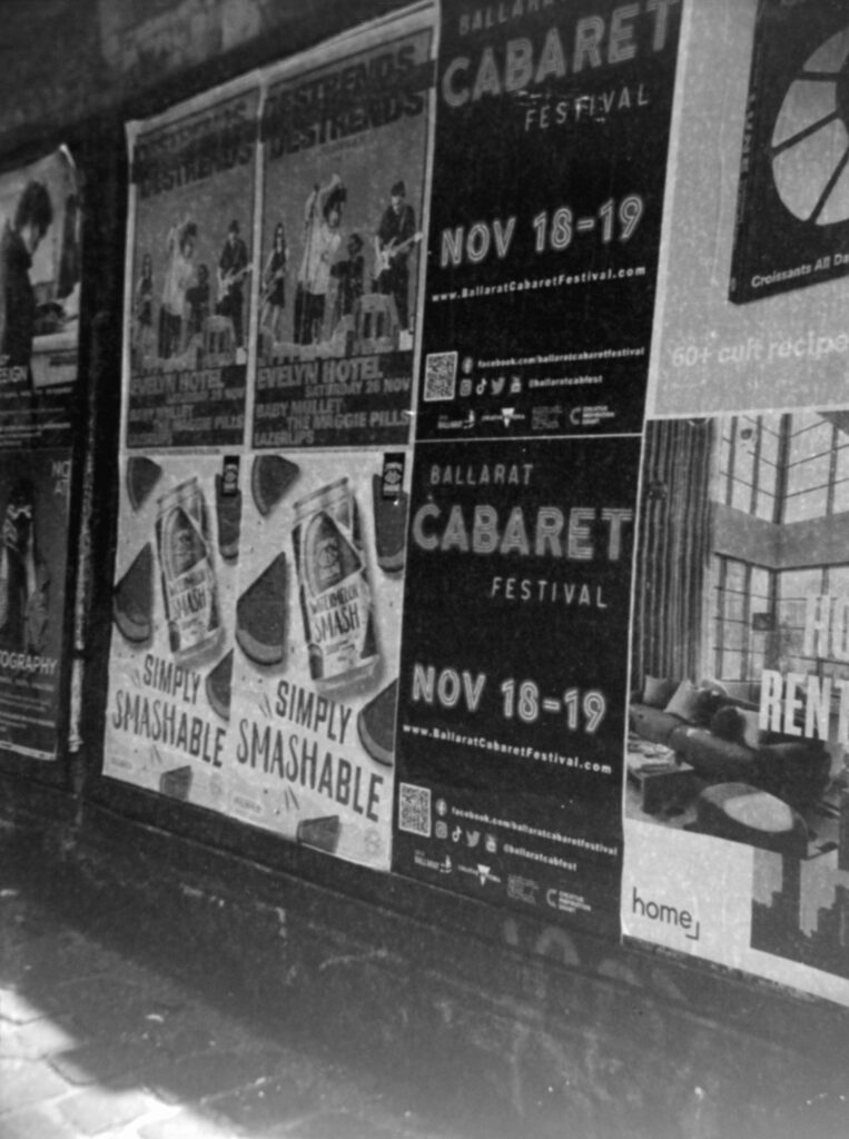Black and white photo of a wall covered in posters advertising drinks, bands, festivals and real estate. There is a bluestone footpath in the foreground