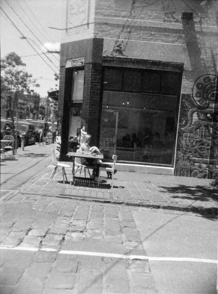 Black and white photo of a woman sitting at a table out the front of a cafe window. She has a dog with her that is partially hidden behind the table. The footpath in the foreground is bluestone and there is street art on the wall next to the cafe's large window.