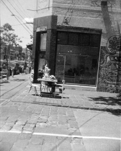 Black and white photo of a woman sitting at a table out the front of a cafe window. She has a dog with her that is partially hidden behind the table. The footpath in the foreground is bluestone and there is street art on the wall next to the cafe's large window.