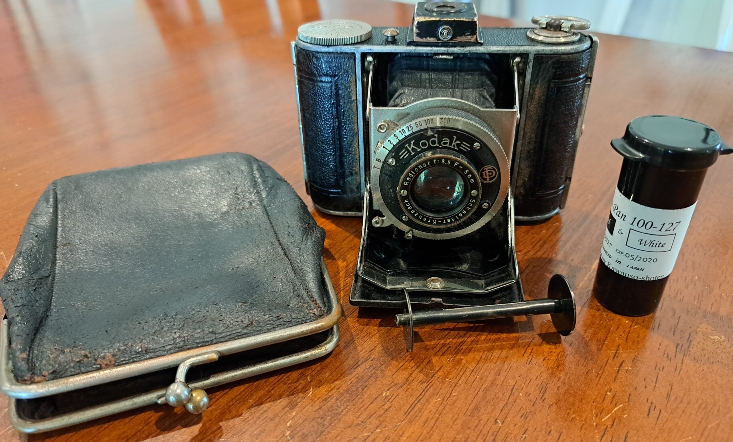 Small vintage fold out camera next to a purse (which it fits into), a roll of ReraPan 100-127 film in a black canister, and a metal 127 take-up spool.