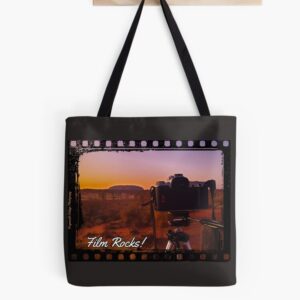 A black tote bag handing on a wooden hook. The bag has an image of photo negative border with an image inside of a film SLR camera pointing at Uluru at sunset. The words "Film Rocks!" are across the bottom. It is signed Frogmouth Cottage Photography