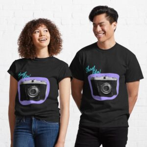 A man and a woman wearing identical T-shirts. The t-shirts are black with a black and white image of a vintage folding camera in a purple border. The words "Shoot Film!" are in blue above the camera.