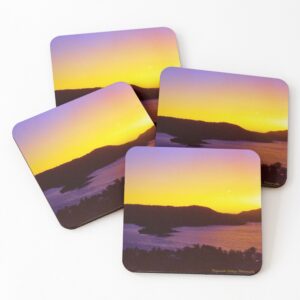 A set of four coasters with a photo of a yellow and purple sunset looking out across the ocean at an island. It is signed Frogmouth Cottage Photography.