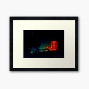 Photo framed in a black frame with a large white border. The photo is taken at night of a neon sign at the top of a building. The sign is in the shape of a left turning arrow with the word "Bar" in the middle. It is signed by Frogmouth Cottage Photograpy.