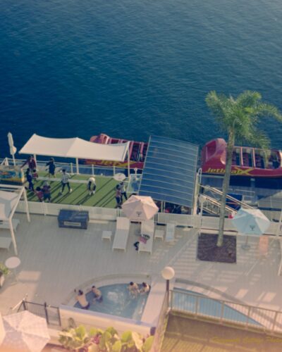 Photo taken from a balcony looking down to a hotel pool area beside a river. The river is a deep blue colour and there are two red speed boats alongside the deck waiting for passengers. There are four people in the spa attached to the pool.