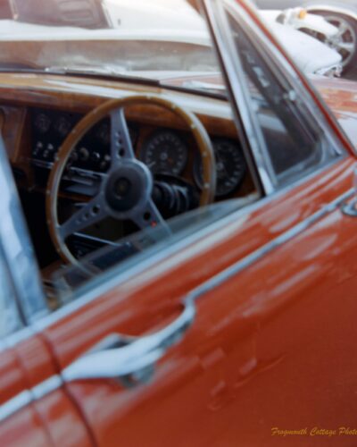 A close up photograph taken looking into the open window of a red car. The front dashboard and steering wheel are made of polished wood.