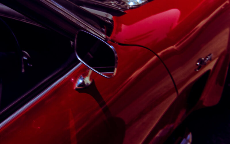 Close up photo of the side door and mirror of ared car. The shining metal of the engine is just visible as the bonnet is up