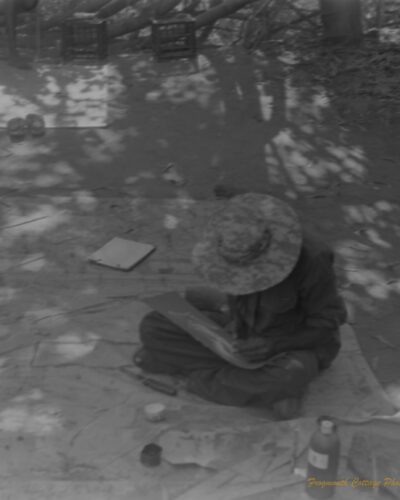 Photo of an indigenous artist sitting cross legged on a canvas. He is paining with a thin piece of reed and looking down at his work.