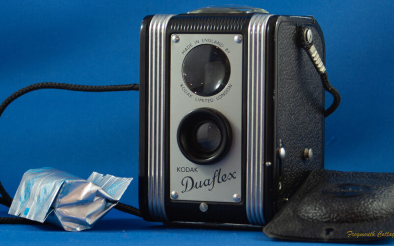 Photograph of a box style camera a plastic cover lying on the right hand side of the image. A cord is attached to either side and is coiled on the left hand side of the image. There is a roll of 120 film lying on the left side of the camera.