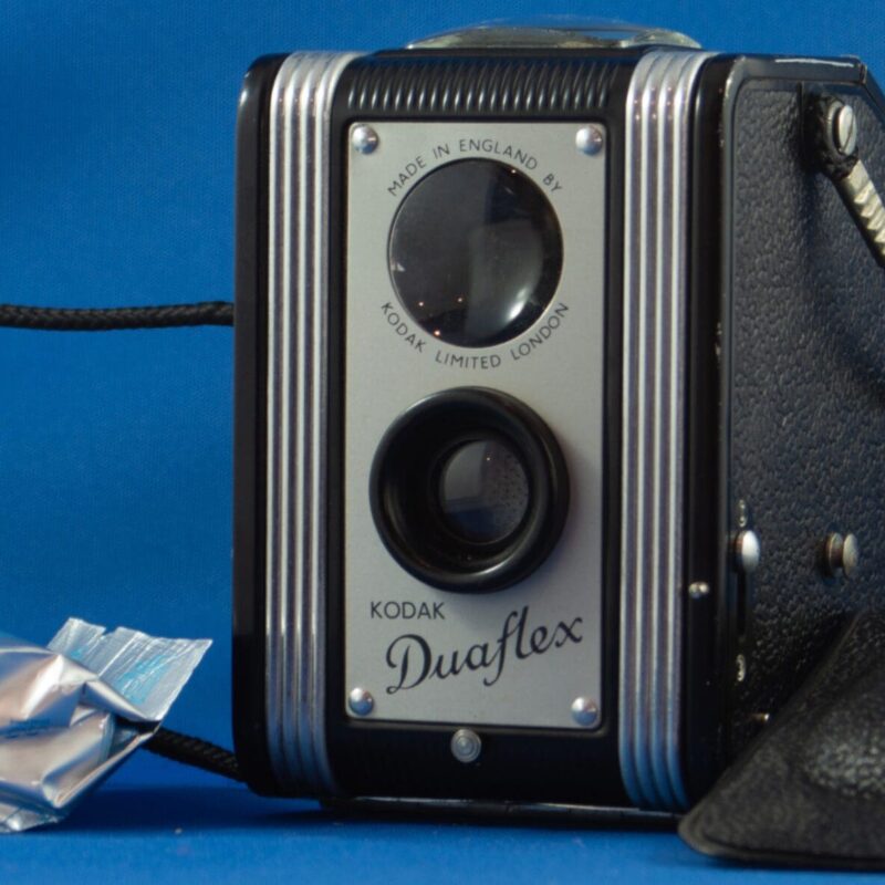 Photograph of a box style camera a plastic cover lying on the right hand side of the image. A cord is attached to either side and is coiled on the left hand side of the image. There is a roll of 120 film lying on the left side of the camera.