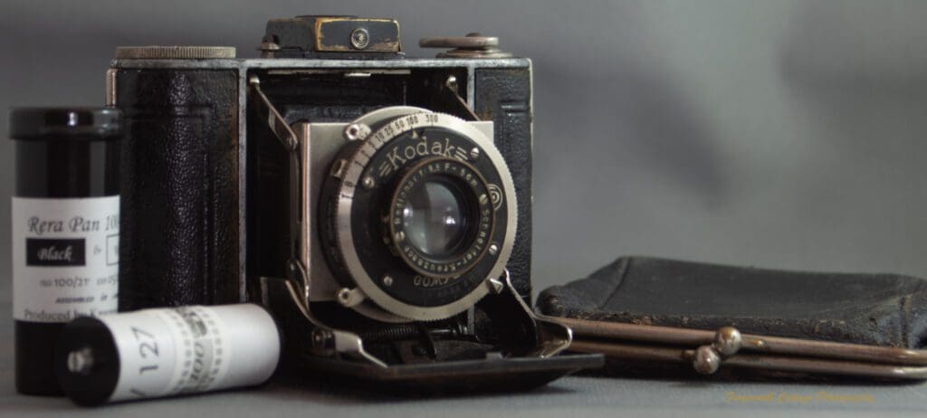 Photograph of a Kodak Vollenda 48 camera - a folding 127 roll film camera. The camera is open with the bellows folded out. There are two rolls of 127 film on the left hand side and a small purse (that the camera fits into) on the other side.