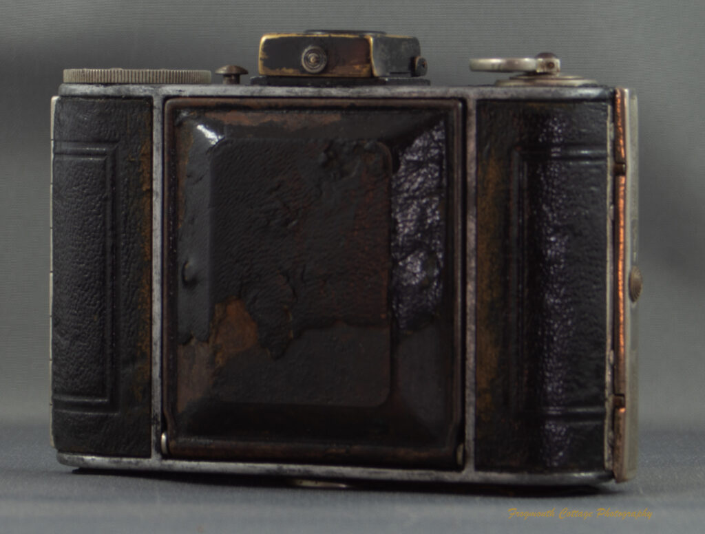 Photograph of a Kodak Vollenda 48 camera - a folding 127 roll film camera. The camera is closed with the bellows folded in. Some of the leatherette on the front of the camera has fallen off and the metal beneath it is rusting.