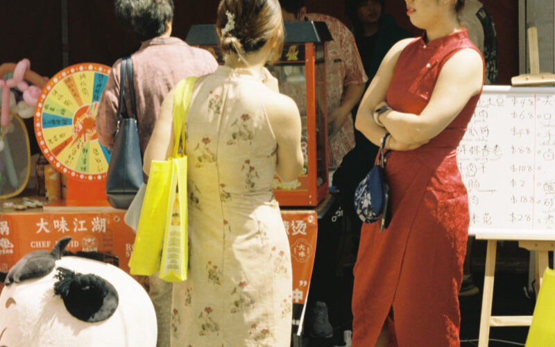 Photo of a woman in red dress and a woman in a cream dress with red flowers standing in the sun in front of a chinese food stall.