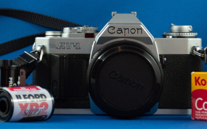 Photograph of a Canon AV-1 SLR camera with a strap and three rolls of film.