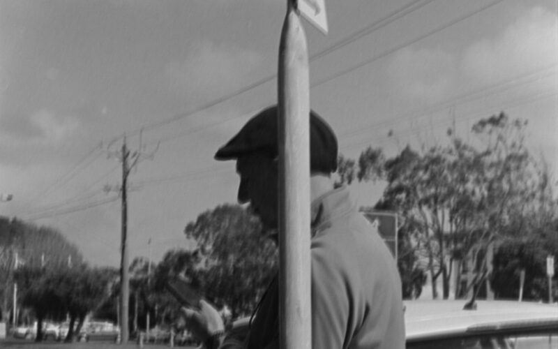 Black and white photo of a man leaning against a sign post. The man is looking at his phone. The sign syas "no standing".