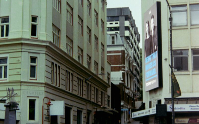 Photograph looking up an alley way of a larger street. There are tall cream coloured buildings eitherside of the alley and two people have just crossed in front of it. There is a large advertising billboard on one of the buildings. The sky is grey and it has been raining.