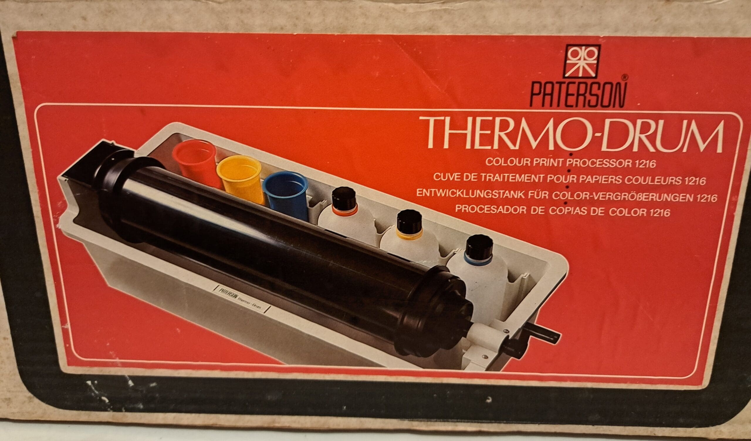 Photograph of the picture on a large box. The background is red with a picture of a paper developing drum, tank, bottles and cups . The label reads: Paterson Thermo-Drum Colour Print Processor 1216.