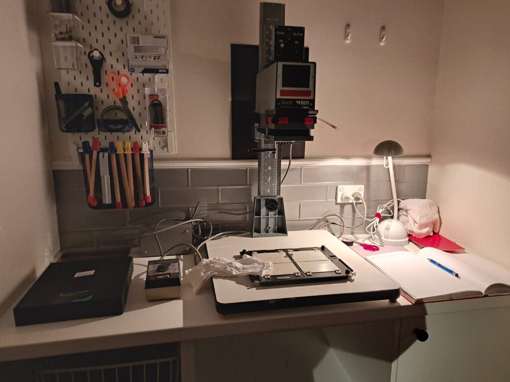 Photograph of a bench with an enlarger in the centre. To the left is a timer and box of paper, and to the right is a desk lamp and notebook and pen. On the wall behind is a pegboard with various tools on it.
