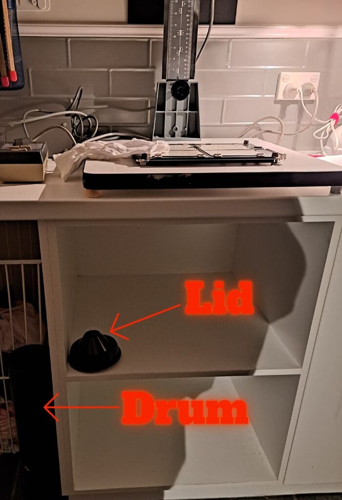 Photograph of the shelving area below a bench with an enlarger on it. Red arrows are pointing at the location of a the drum and lid placed below the bench.