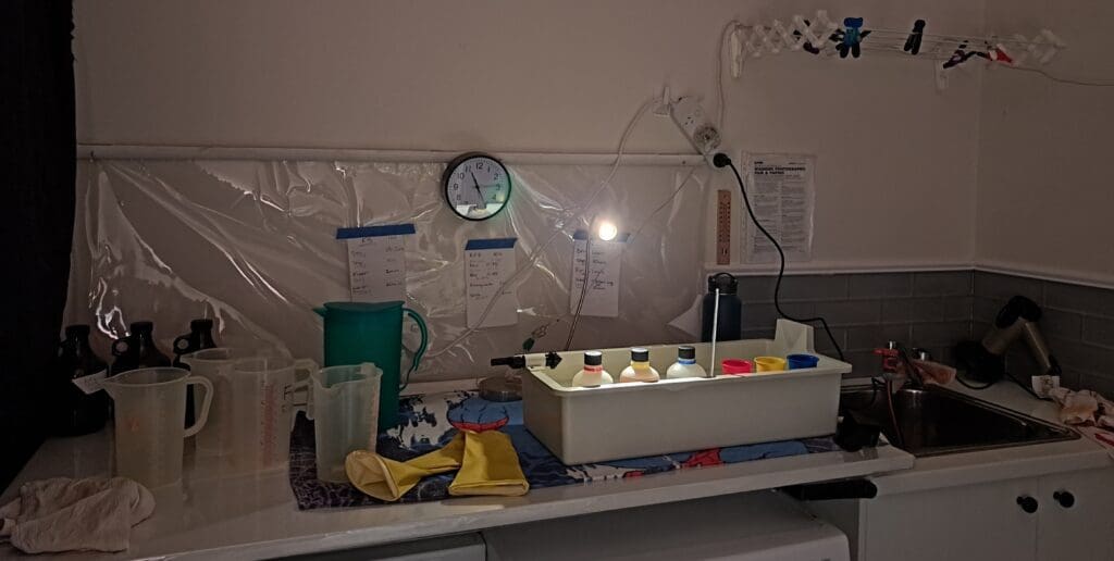 Photograph of a bench lit by a desk lap with brown glass jugs and plastic jugs on the left hand side. Next to this is a colour processing water batch with bottles and cups sitting inside. On the right hand side is a sink and on the wall behind are note pages and a clock.