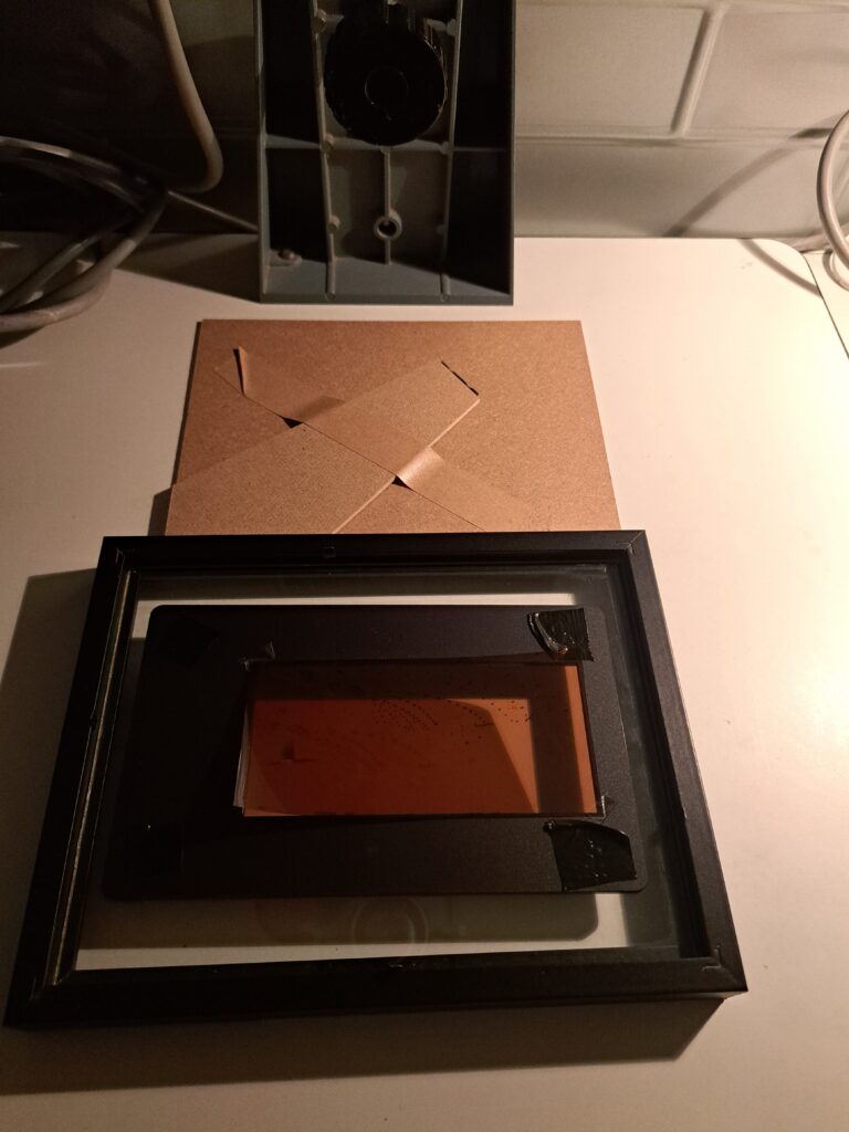 A photo frame lying glass side down, with the back removed and placed behind it. A negative has been placed on the glass and a black plastic mask placed under it to create a border and cover the edge markings on the negative. Small pieces of tape hold the negative to the mask.