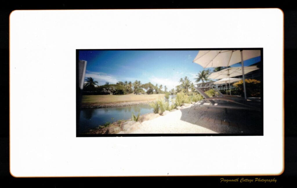 Colour photograph taken on a pinhole camera looking across a stream to a grassy area and trees in the background. On the right hand side are lounge chairs and umbrellas with apartment buildings in the background. The image has a large white border and a smaller black border around it.