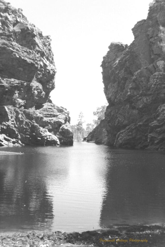 Black and white photograph of a large watering hole between two large rocky outcrops.
