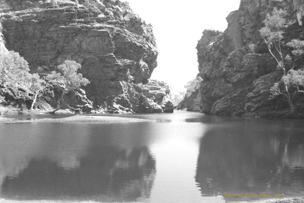 Black and white photograph of a large watering hole between two large rocky outcrops.