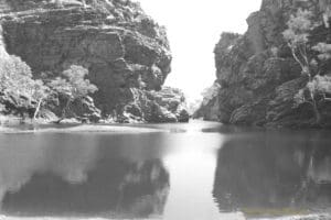 Black and white photograph of a large watering hole between two large rocky outcrops.