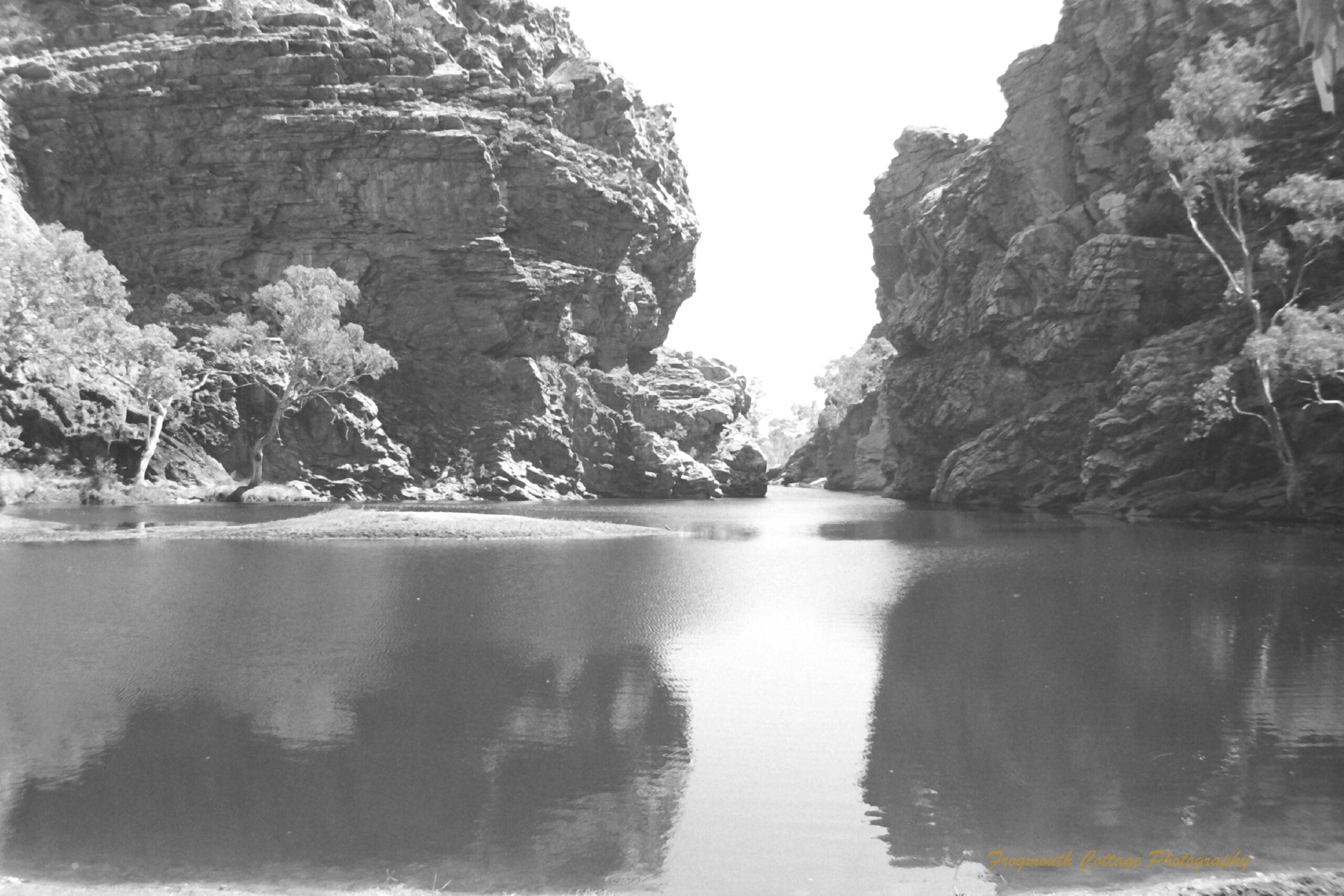 Black and white photograph of a large watering hole between two large rocky outcrops.