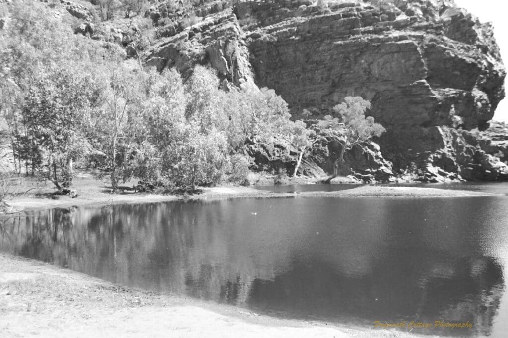 Black and white photograph of a large watering hole between two large rocky outcrops.
